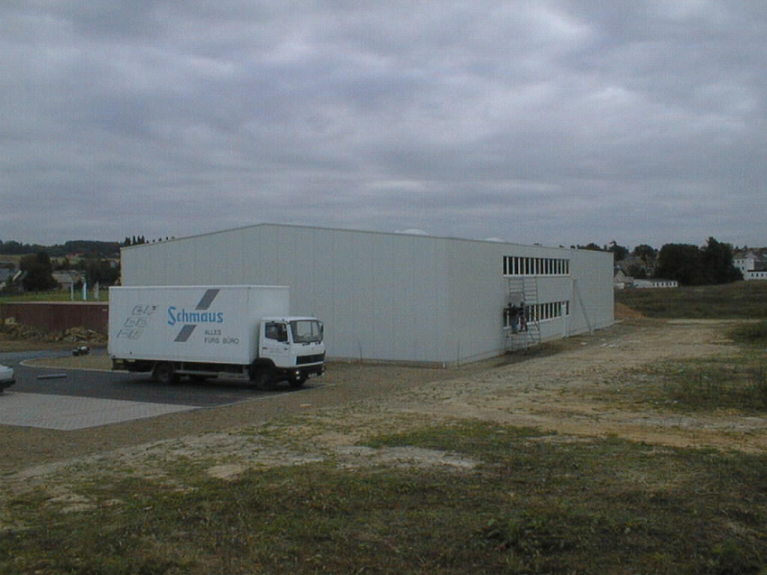 Von einer Anhöhe fotografiert: Außenansicht der Logistikhalle nach dem Schließen der äußeren Gebäudehülle. Der Lkw mit "Schmaus"-Firmenlogo steht im Vordergrund auf dem ebenfalls neuen Parkplatz. Die Geländeflächen sind erdig eingeebnet und noch ohne Gras oder Begrünung. Auf einem Baugerüst hantieren noch Arbeiter am Fensterband. 